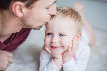 Blond-haired blue-eyed baby girl and young dad in the home interior. Dad kisses his little daughter.