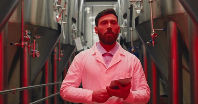 Scared male worker in white lab coat holding tablet and walking through rows of steel brewing vats. Alert signals and red lights at brewery factory. Concept of emergency situation