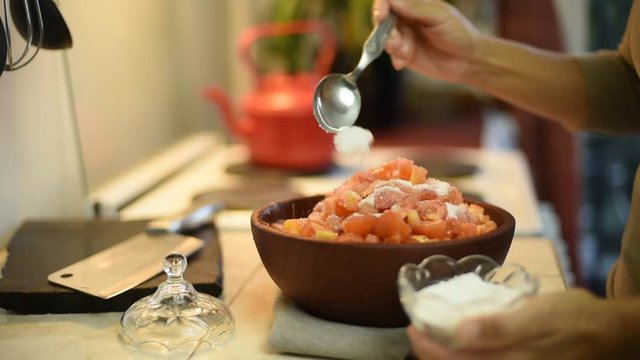 manos poniendo azucar sobre tomates cortados en un cuenco de madera sobre mesada de una cocina 