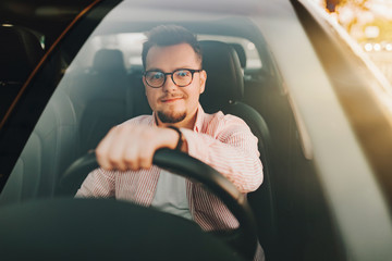 A young europen beautiful smiling man wearing glasses driving a car in the sity on sunset. View through the windshield