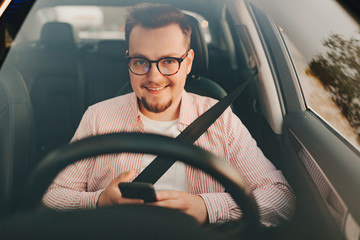 Obraz premium A car driver sitting in a car with seat belt and smartphone. View .through the windshield of a stylish smiling man in glasses