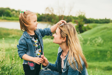 A little blonde girl walks with her mother in nature. The girl adjusts the glasses on her mother's head. Mother and daughter dressed in denim jackets walk among the rural meadows near the pond