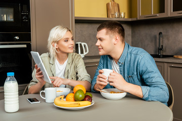 woman showing digital tablet to husband while having breakfast in kitchen