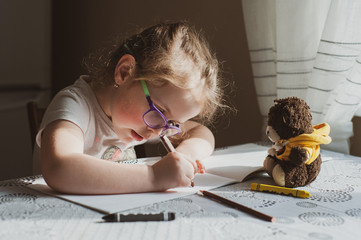 A little girl with glasses draws a portrait of a soft teddy bear, which lies in front of her. Idea...