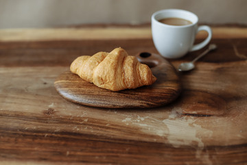 Fresh croissant lies on a wooden board with a cup of coffee on a wooden table.