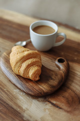 Fresh croissant lies on a wooden board with a cup of coffee on a wooden table.