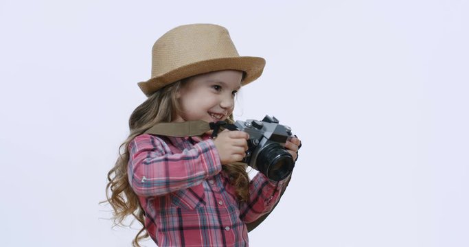 Nice pretty joyful Caucsian small girl with fair hair and in hat taking a photo with photocamera and smiling cheerfully. White wall background.