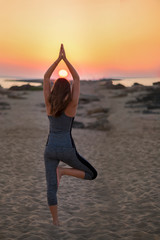 Fototapeta na wymiar Young sporty fit woman is doing Hatha yoga asana Vrikshasana tree pose on beach on sunset, Yoga outdoors