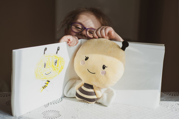A little girl with glasses draws a portrait of a soft toy bee that lies in front of her. Idea for quarantining a child © Kiryakova Anna