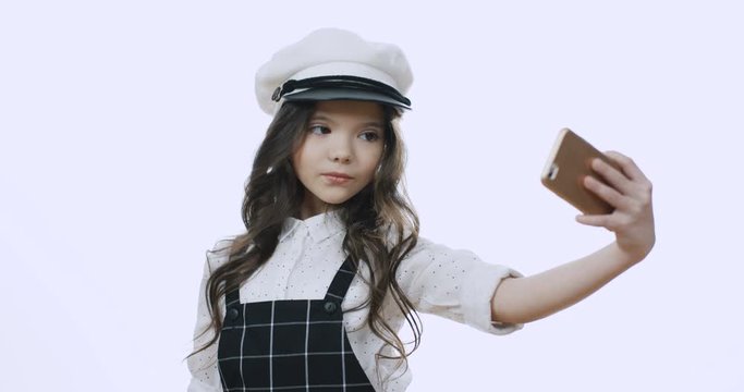 Cute Caucasian stylish and charming small girl in the hat taking selfie photo on the smartphone camera and posing like a model. White wall background.