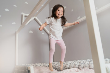 Happy little girl wearing white pajamas jumping on bed in white bedroom. Cute girl having fun while...