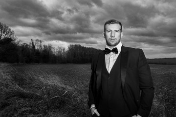 Handsome man wearing tuxedo standing with fields and stormy sky behind him.