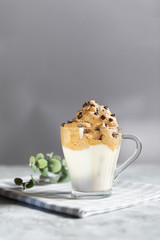 Dalgona frothy coffee in a transparent glass cup decorated with chocolate drops, trend korean drink made with milk and coffee foam on grey background