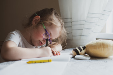 A little girl with glasses draws a portrait of a soft toy bee that lies in front of her. Idea for...
