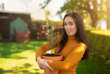 Beautiful dark haired smiling woman holding pot with plant while standing in the garden