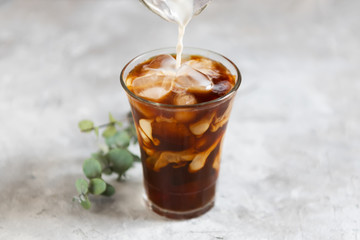 Milk is pouring into a tall glass with brew black coffee with pieces of ice on a gray dark background, close-up. Ice summer fresh coffee concept