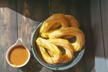 Churros with sugar and caramel sauce on wooden rustic table. Traditional Spanish or Mexican dessert. Homemade cookies, beautiful morning light at kitchen, sunny day. Top view, copy space.
