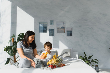 mom with son in pajamas eating fruit on bed. portrait happy, smiling, joyful family, mothen and son having breakfast in bed, surprise on mom day. Positive human emotions