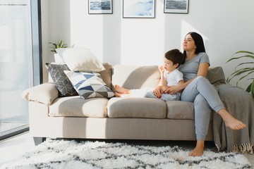 Cheerful mother sitting on couch in cozy light living room holds on lap tickles of little son enjoy...