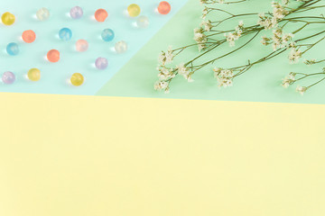 Bright color block paper background (yellow, light blue and green) with some multicolored pastel beads and white flowers of gypsophila. Copy space. Space for text. Flat lay, top view.