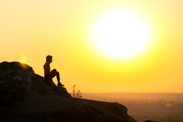 Fototapeta premium Silhouette of a woman hiker sitting alone on big stone at sunset in mountains. Female tourist on high rock in evening nature. Tourism, traveling and healthy lifestyle concept.
