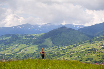Back view of slim young woman standing on grassy valley on background of green mountains on sunny summer day.