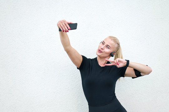 Young attractive sporty girl wearing black sportswear listening to music and stretching on white wall background. Concept active and healthy life.