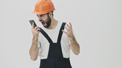 Screaming stressed angry young bearded man builder isolated over white wall background in helmet talking by mobile phone