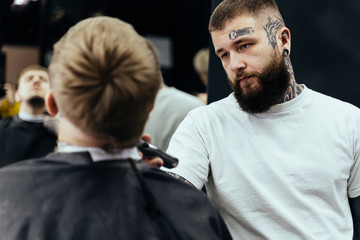 Tattooed Barber trimming bearded man with shaving machine in barbershop. Hairstyling process. Close-up of a Hairstylist cutting the beard of a bearded male.
