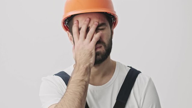 Tired yawning young bearded man builder isolated over white wall background in helmet