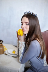 A brunette in a gray dress embroidered with lace holds a Ranunculus flower near her face. Queen. A gorgeous girl with her eyes closed.