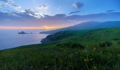 Fototapeta na wymiar Green hills and steep rocky shores by the ocean. View of the rugged islands and peninsulas at sunset. In the foreground is green grass.