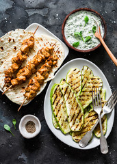 Chicken skewers, grilled zucchini, tortillas and tzadziki sauce - delicious greek style lunch on a dark background, top view
