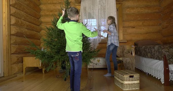 Mother with two children dresses up Christmas tree for the holiday. In the interior of the village house, a mother with children amicably take out Christmas toys and hang them on a green fluffy tree.