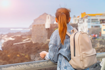Fototapeta na wymiar Redhead girl with a backpack admire ocean and architecture of Garachico, Tenerife on a sunny day. Travel concept. Tourist woman explore location with a small rucksack.