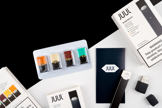 Juul vaping device and juul flavoured nicotine pods, next to usb charger and packages with warning tag. On black and white background. Futuristic cigarettes and technology concept.