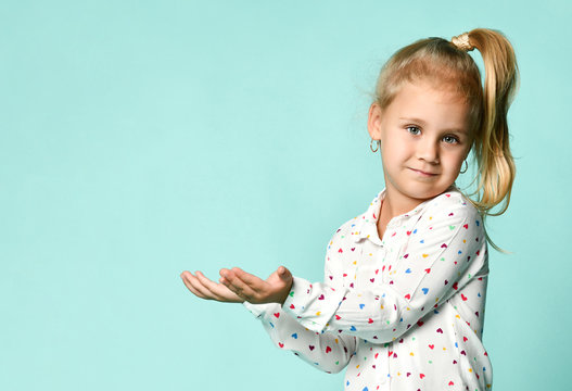 Little Blonde Girl With Ponytail, In Shirt With Hearts Print. She Smiling, Holding Something Posing On Blue Background. Close Up