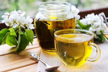 a clear Cup of tea and liquid honey in a jar on a light wooden table by the window. next to the white flowers of a plum or Apple tree