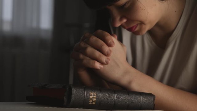 A woman prays with her hands crossed over a Bible for God's blessing in a dark room.