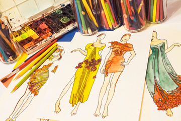 Workplace of a women's costume designer. Paints, colored pencils, and brushes are scattered on the table. In the center are artistic drawings of model girls in evening dresses.