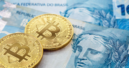 electronic bitcoin coins, with a hundred dollar bill in the background. Devaluation of the Brazilian real against the decentralized currency.