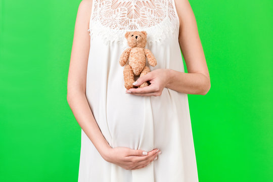 Cropped image of teddy bear in hand against pregnant woman's belly in white dress at green background. Waiting for a childbirth. Copy space