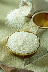 Uncooked dry Thai jasmine rice in bowl is isolated and burlap bag with lid of bowl, wooden ladle put on brown fabric, warm tone and vertical view.