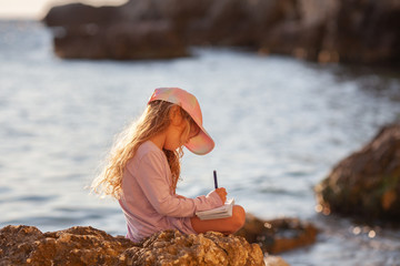 Happy pretty girl walks along the sea coast against the background of the sea, from behind a beautiful landscape. The child draws in a notebook