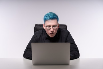 Fashionable man with blue hair seriously working on his laptop, studio photo with white background