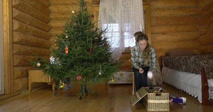 Mother with two children dresses up Christmas tree for the holiday. In the interior of the village house, a mother with children amicably take out Christmas toys and hang them on a green fluffy tree.