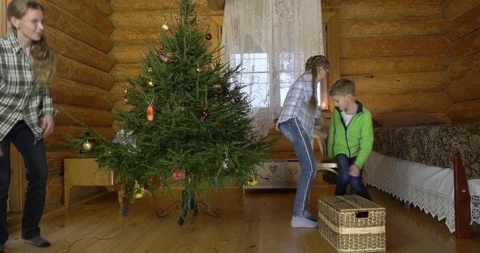 Mother with two children dresses up Christmas tree for the holiday. In the interior of the village house, a mother with children amicably take out Christmas toys and hang them on a green fluffy tree.