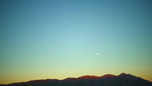 Beautiful stable shot of the moon over the orange mountains with the blue sky as a background in dusk time
