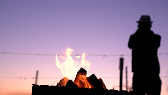 Silhouette of a man who stands back to the camera and recording video of a beautiful colorful sunset on the phine, a fire burns in the grill
