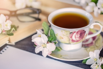 desktop: a cup of tea, a book, glasses, a sheet of Notepad and branches of flowers. the concept of a pleasant time: tea, book, flowers. background with a cup of tea, a book, and flowers.